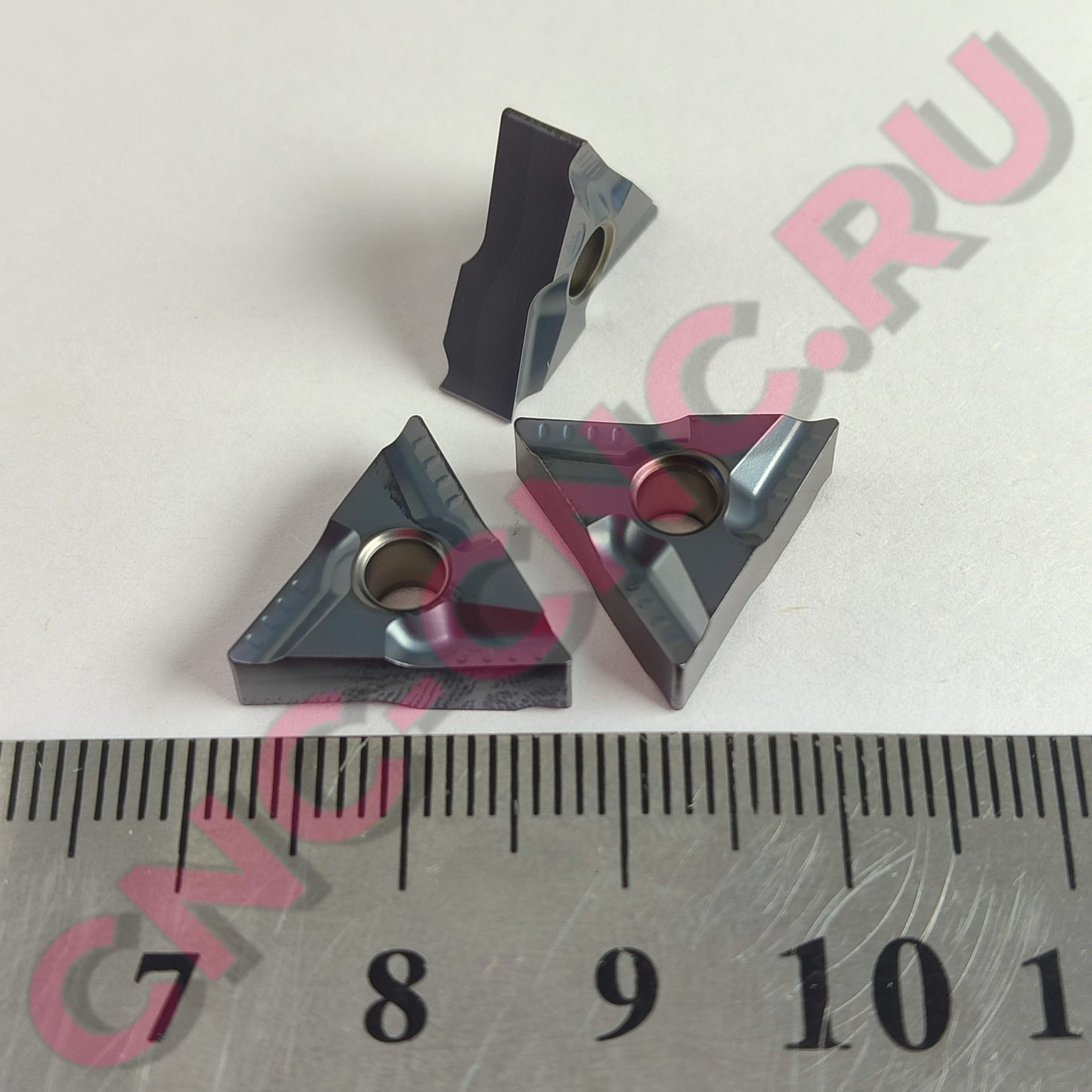 Close-up of glossy gray angular plastic pieces with circular holes scattered on a ruler marked in centimeters. The pieces have pink translucent accents and sit on a light surface.