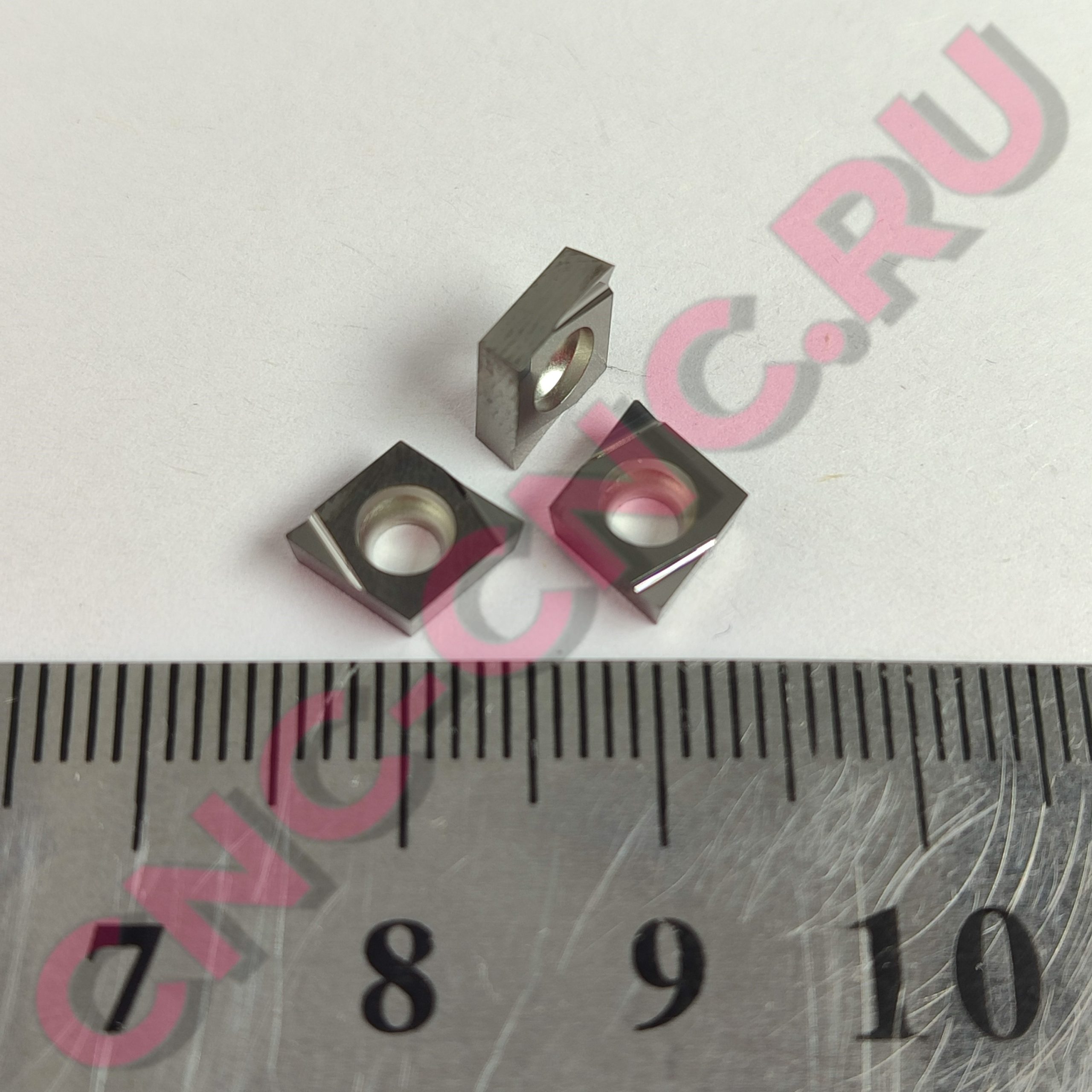 Close-up of a metal ruler with inch marks and black numbers, overlaid by pink watermark text and metal blocks obscuring part of the scale.