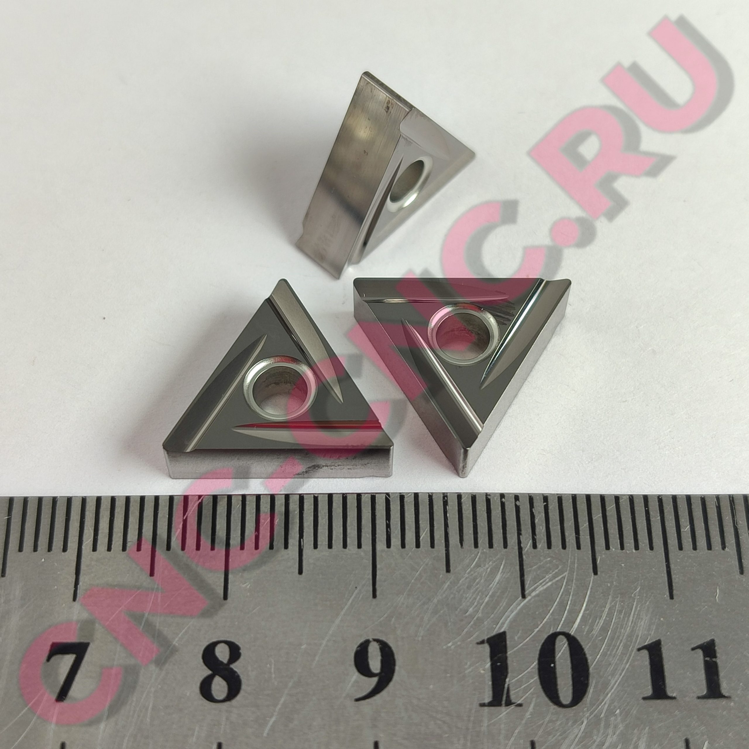 Close-up of several shiny, triangular metal blocks with circular holes, arranged on a ruler-scale background.